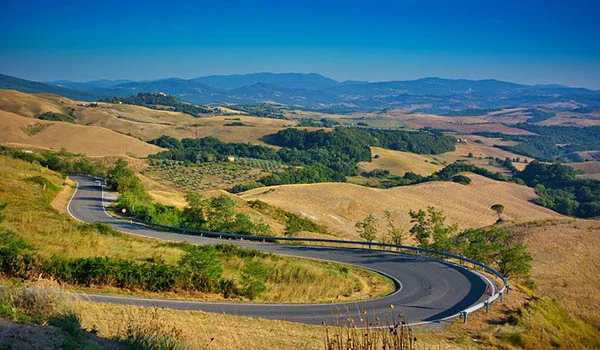 Estrada sinuosa atravessa colinas douradas e onduladas da Toscana, com vinhedos, árvores e montanhas ao fundo.