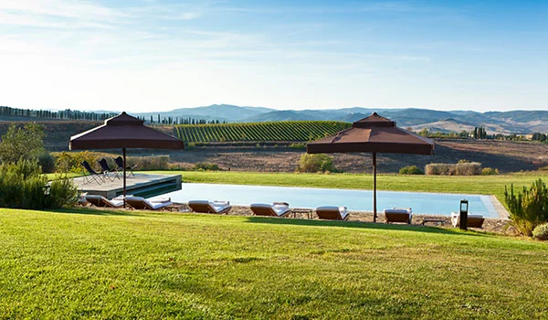 Piscina ao ar livre com espreguiçadeiras e guarda-sóis, cercada por gramados e vinhedos nas paisagens da Toscana.