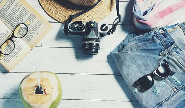 Imagem estilosa de verão com câmera, chapéu, óculos escuros, um livro e uma bebida refrescante em ambiente descontraído. Dicas para presente quem gosta de viajar.