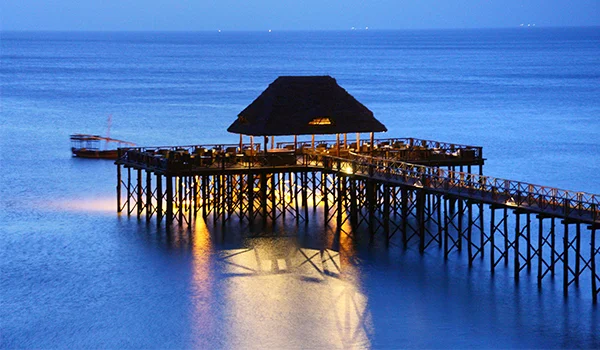 Píer de madeira sobre o mar ao entardecer, com um restaurante coberto por um grande teto de palha e luzes refletidas na água.