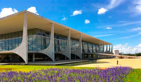 Fachada do Palácio do Planalto com arquitetura moderna, grande estrutura de concreto, vidro e um jardim de lavanda na frente, sob céu azul com nuvens. Lua de Mel em Brasília.