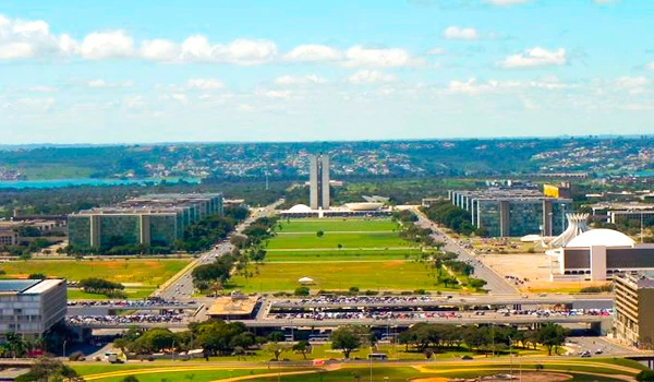 Vista aérea do Plano Piloto com seus amplos gramados, edifícios modernos e avenida principal ao centro, sob céu parcialmente nublado. Lua de Mel em Brasília.