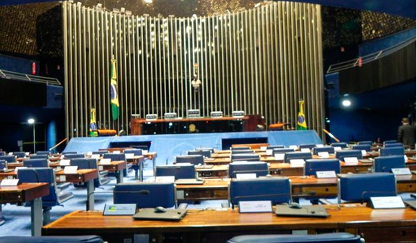 Sessão de votação na Câmara dos Deputados do Brasil, com assentos azuis e microfones em cada bancada, decorada com bandeiras brasileiras e cenário formal ao fundo.