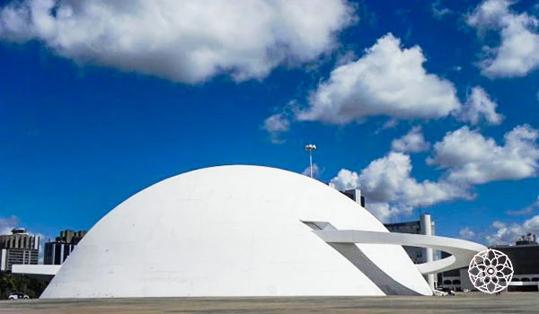 Imagem da Ópera de Arame em Curitiba, um espaço cultural e de eventos com arquitetura moderna e estrutura única de tubos de aço, sob céu azul com nuvens.