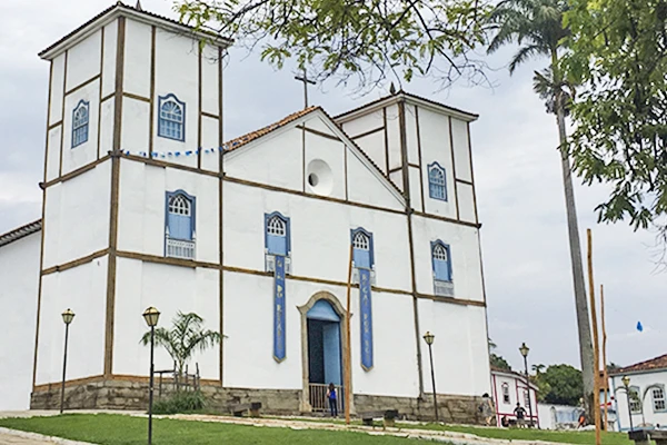 Igreja histórica com arquitetura simples, fachada branca com detalhes azuis, situada em um ambiente com gramado bem cuidado e árvores ao redor. Atrações do Planalto Central.