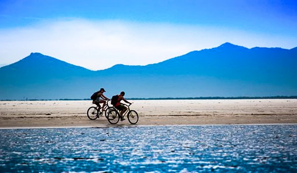 Duas pessoas pedalando suas bicicletas na beira de um lago em um cenário ao entardecer ou amanhecer, com montanhas ao fundo e céu claro, promovendo lazer e contato com a natureza.