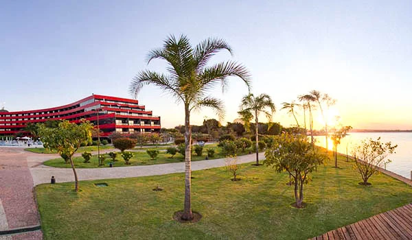 Imagem de um hotel moderno ao lado de uma praia ao pôr do sol, com árvores, gramados bem cuidados e um ambiente tranquilo. Ideal para a Lua de Mel em Brasília.