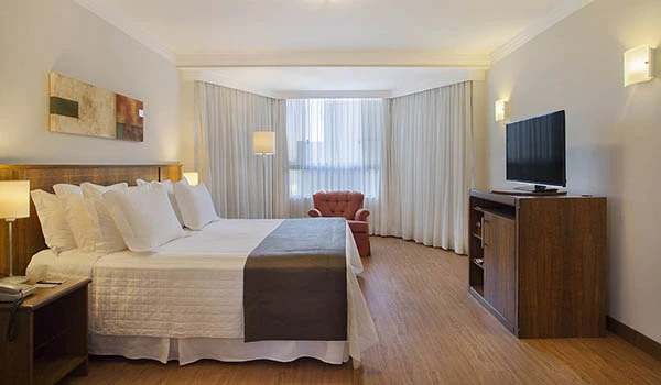 Quarto de hotel espaçoso com cama de casal, poltrona vermelha ao fundo, televisão na bancada de madeira e cortinas brancas em ambiente bem iluminado.