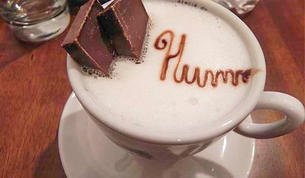 Imagem de uma xícara de café com a palavra 'Happy' feita com cacau em pó ou canela, decorada com dois pedaços de chocolate ao leite por cima, criando uma apresentação deliciosa e acolhedora