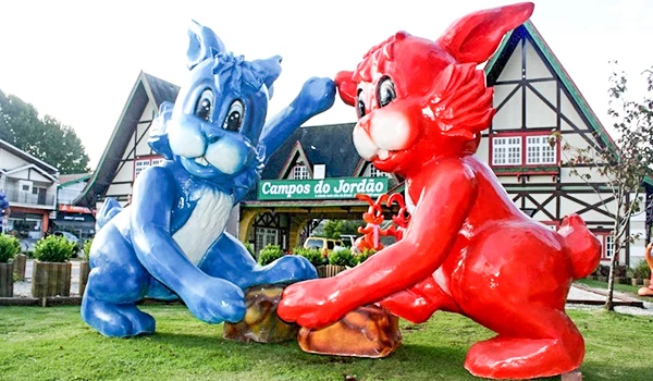 Estátuas de coelho azul e coelho vermelho em frente ao Campos do Jordão, com casas de arquitetura típica na região, ideal para fotos e lazer.