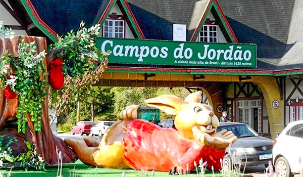 Escultura de coelho gigante decorativo na entrada de Campos do Jordão, com elementos natalinos e ambiente ensolarado, destacando atrações turísticas do destino.
