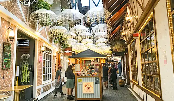 Imagem de um shopping ao ar livre com decoração de guarda-chuvas coloridos e iluminação charmosa, onde pessoas fazem compras e socializam.