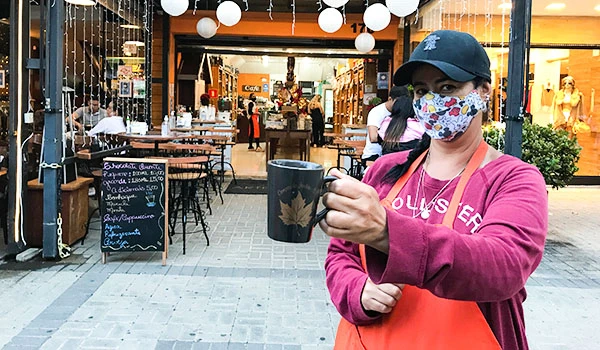 Mulher usando máscara de proteção com estampa floral, segurando uma xícara de café em uma rua movimentada de shopping, com lojas ao fundo ao ar livre.