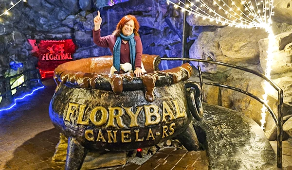 Deusa Rodrigues dentro de uma enorme panela de cobre na atração Florybal em Canelinhas, Santa Catarina, com iluminação de festa ao fundo, promovendo turismo local.