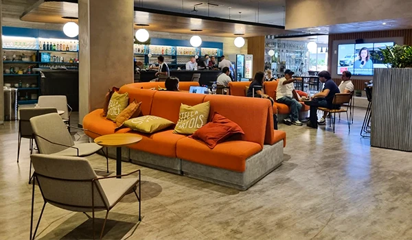 Foto do Novotel de Belo Horizonte, um ambiente descontraído de um café com sofás laranjas, mesas e pessoas trabalhando em laptops.