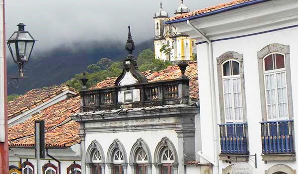 Imagem da arquitetura colonial de Ouro Preto, mostrando telhados de barro, janelas com detalhes em azul e o famoso cenário das montanhas ao fundo.