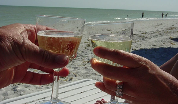 Casal brindando com taças na praia, refletindo um momento descontraído e alegre sob o sol. Lugares para comemorar aniversário de casamento.