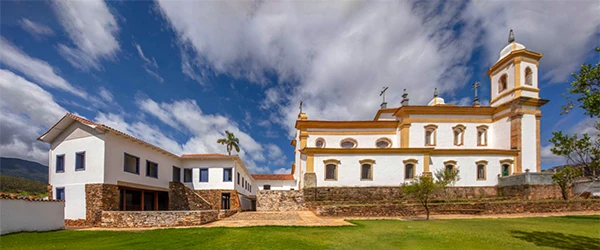 Foto de uma igreja histórica e edifício anexo em um dia claro, com céu azul e nuvens em Mariana, para Lua de Mel em Minas Gerais.