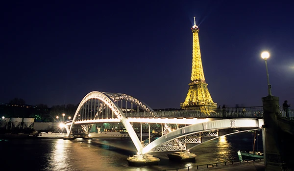 Vista noturna da Torre Eiffel iluminada, com uma ponte em primeiro plano, capturando a beleza de Paris à noite.