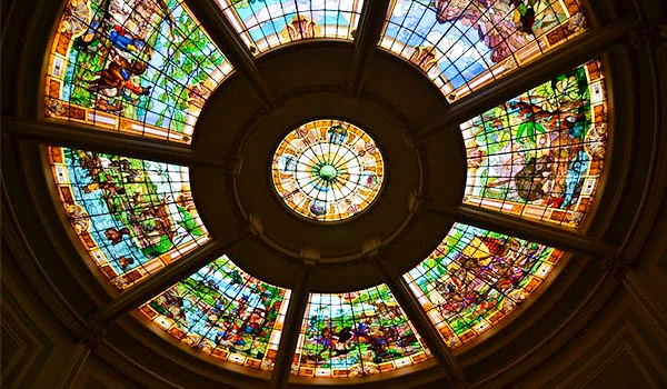 Foto de uma claraboia com vitrais coloridos no Grande Hotel de Araxá, mostrando uma magnífica cúpula decorativa com detalhes em vidro. A iluminação natural realça as cores vibrantes e a arte dos painéis, criando uma atmosfera única e encantadora.