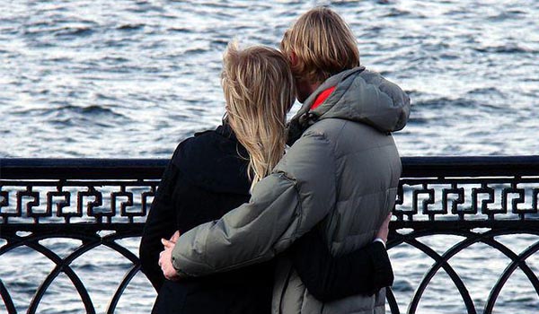 Foto de um casal de costas, abraçados e de frente uma ponte gradeada de um rio. 