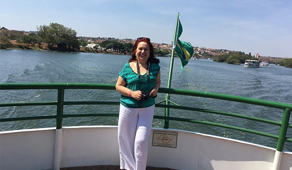 Deusa Rodrigues posando no barco de Barra Bonita com vista para o rio, segurando uma câmera e com uma bandeira ao fundo.
