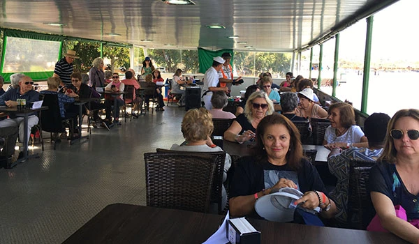 Imagem de um restaurante com várias pessoas sentadas à mesa, desfrutando de uma refeição em ambiente ao ar livre sob cobertura, com paredes de vidro e iluminação natural