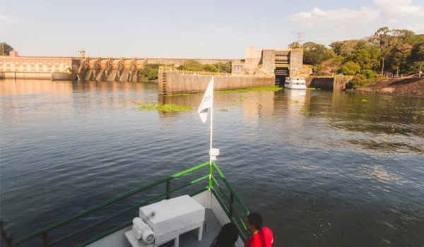 Embarcação de passeio navegando por um rio com uma estrutura de represa ao fundo, céu claro e cenário natural ao redor na cidade. Barra Bonita passeio de barco.