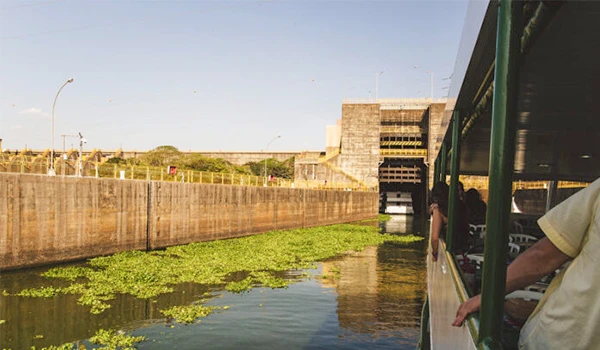 Imagem de uma embarcação em um canal de água, com estrutura de cimento ao fundo, pessoas a bordo e vegetação ao redor, sob céu claro. Barra Bonita passeio de barco.