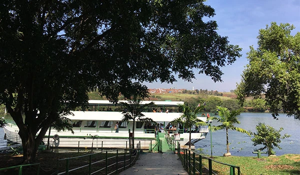 Paisagem com um barco grande ao lado de um rio, cercado por árvores verdes e céu azul, em uma cena ao ar livre. Barra Bonita passeio de barco
