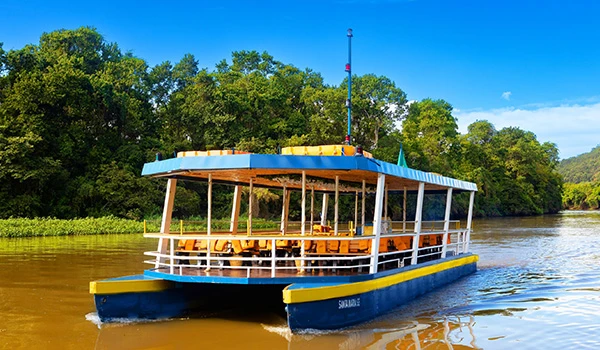 Fotografia de um barco a motor navegando em um rio cercado por vegetação verde exuberante, com céu azul claro, no Parque Três Pescadores em Aparecida. Um dos lugares baratos para Lua de Mel em SP.