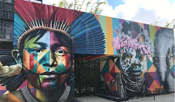 Mural colorido em uma parede urbana, com retratos artísticos de pessoas com pinturas faciais geométricas e adornos tradicionais. À esquerda, destaca-se um rosto com cocar de penas vermelhas e azuis; à direita, uma figura com flores rosas na cabeça, pinturas faciais e colares. Ao fundo, formas geométricas e cores vibrantes compõem a obra, que celebra a diversidade cultural e a identidade dos povos indígenas.