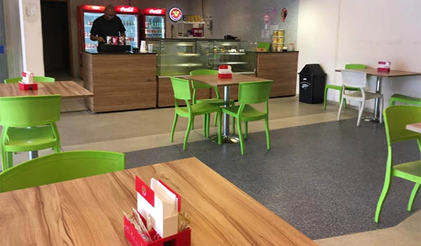 Imagem de uma área de lanchonete com mesas de madeira e cadeiras verdes, ambiente limpo e bem organizado, ideal para refeições rápidas.