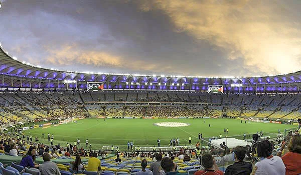 Estádio lotado antes de um jogo de futebol com torcedores animados, céu parcialmente nublado e iluminação especial. Visita ao Maracanã.