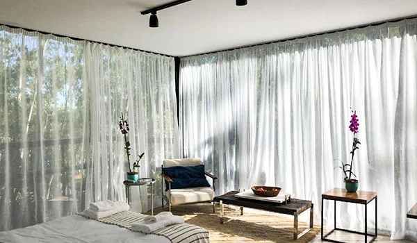 Sala de estar ampla e iluminada, com grandes janelas cobertas por cortinas brancas translúcidas, poltrona, mesa lateral com vaso de planta, sofá claro, mesa de centro de madeira e duas orquídeas decorativas. A luz natural entra pelas cortinas e cria um ambiente aconchegante e integrado à paisagem externa.
