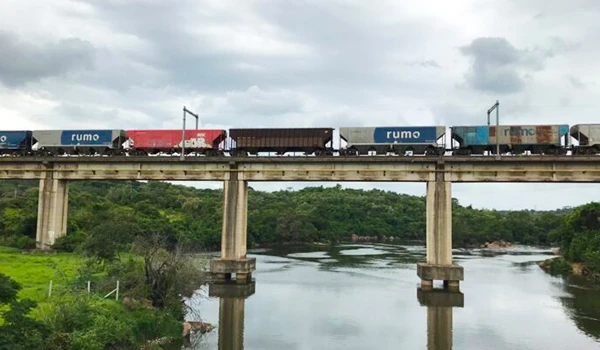 Imagem de um trem de carga passando sobre uma ponte em uma área com rios e vegetação verde sob céu nublado, simbolizando transporte e movimentação logística.