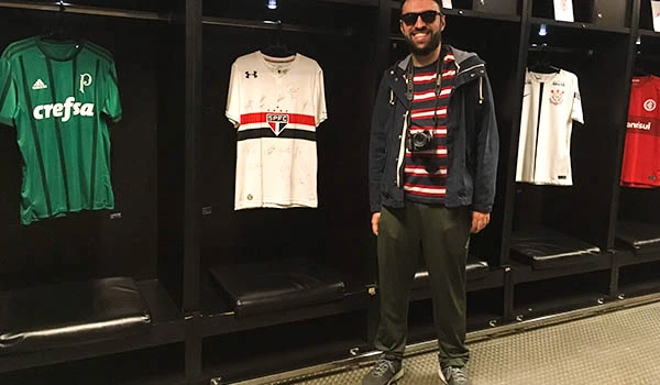 Homem sorridente posando em frente a uma parede com camisas de times de futebol. Visita ao Maracanã.