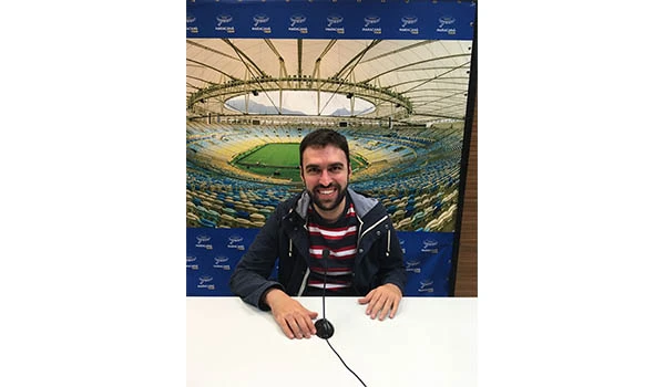 Homem sorridente sentado em mesa na cobertura de um estádio, com fundo de arquibancadas e teto de vidro, promovendo evento esportivo. Visita ao Maracanã.