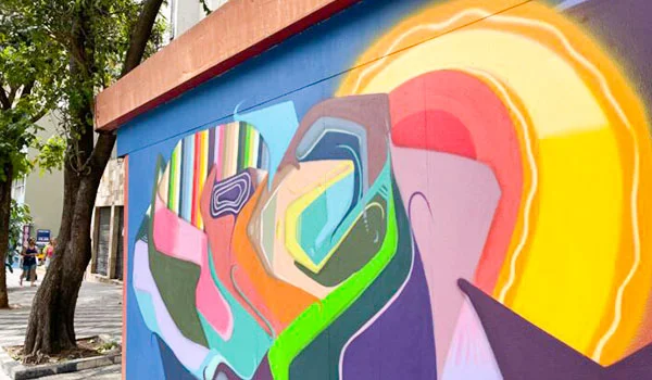 Mural colorido de grafite em uma parede urbana, com formas abstratas e geométricas em tons vibrantes de amarelo, laranja, rosa, azul, verde e roxo. À esquerda, uma árvore sombreia a calçada, por onde circulam pedestres.