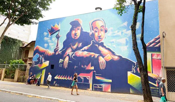 Mural colorido em uma fachada de prédio, retratando três figuras humanas em estilo artístico, com cores vibrantes e elementos geométricos. Árvores emolduram a obra, enquanto pedestres caminham pela calçada em frente ao grafite.