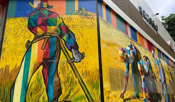 Mural colorido em uma parede externa, retratando trabalhadores rurais em uma plantação de trigo, com figuras humanas pintadas em cores vibrantes e traços expressivos.
