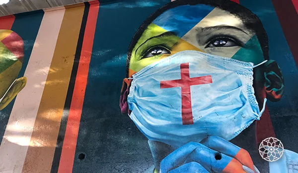 Mural de grafite retrata o rosto de uma pessoa usando máscara cirúrgica azul com uma cruz vermelha, sobre um fundo colorido com formas geométricas e tons de azul, verde, amarelo e vermelho.