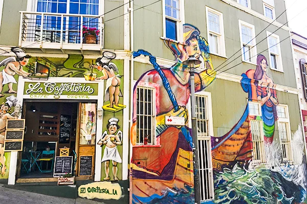 Fachada de um edifício em Valparaíso, no Chile, decorada com grandes murais coloridos que retratam figuras humanas em estilo artístico. À esquerda, funciona a cafeteria La Caffetteria, com mesas e cadeiras na entrada. As pinturas cobrem as paredes do térreo, integrando-se à arquitetura do local, enquanto as janelas, sacadas e os fios elétricos completam a cena urbana.