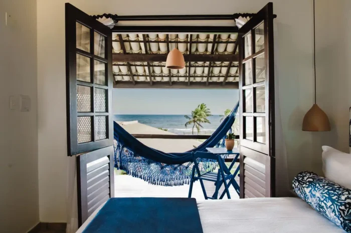 Quarto com cama de casal, mesa de apoio e janela aberta com rede azul e vista para o mar ao fundo, em ambiente tropical com plantas em vaso. Lua de Mel em Salvador.
