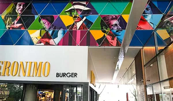 Fachada de uma lanchonete com letreiro “Geronimo Burger”, decorada com um grande mural colorido de rostos humanos em formas geométricas triangulares.