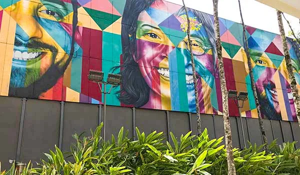 Mural colorido em uma fachada urbana, com grandes retratos de rostos humanos pintados em estilo geométrico, utilizando cores vibrantes como vermelho, amarelo, azul, verde e rosa. Na parte inferior, há vegetação tropical e, à frente do mural, troncos de palmeiras.