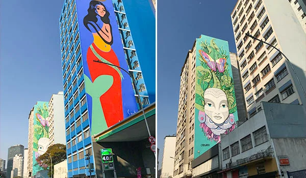 Fotografia de dois prédios altos na Rua da Consolação, em São Paulo, com grandes murais coloridos de arte urbana nas fachadas. À esquerda, uma mulher de cabelos pretos, blusa amarela e saia vermelha; à direita, um rosto feminino com adornos florais, borboletas e folhagens. O céu está azul e sem nuvens.