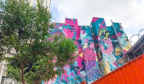 Mural colorido de grafite em fachadas de prédios na região do Minhocão, no centro de São Paulo, com formas abstratas em tons vibrantes de rosa, azul, verde e amarelo. Árvores ocupam o lado esquerdo da imagem, enquanto uma barreira laranja com arame farpado aparece na parte inferior direita.