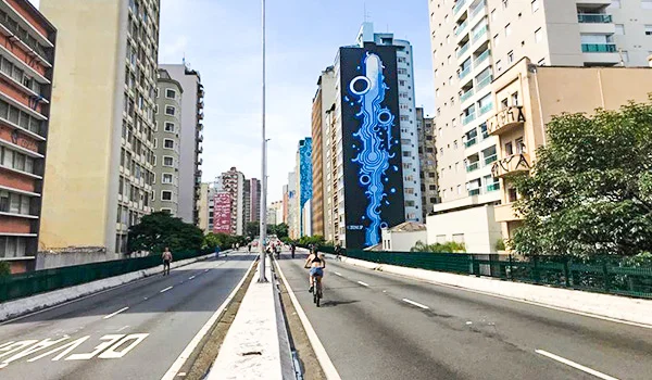 Vista de uma avenida urbana em São Paulo, com prédios altos, ciclistas e pedestres circulando pela via. Ao centro, destaca-se um edifício com um grande mural de grafite azul em estilo futurista, com formas abstratas e elementos que lembram circuitos eletrônicos.