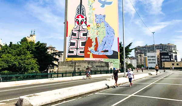 Mural colorido com figuras de animais e desenhos geométricos em um grande painel na lateral de um edifício, em uma avenida urbana de São Paulo. Ao redor, há árvores, prédios, pedestres e ciclistas sob um céu azul com nuvens.